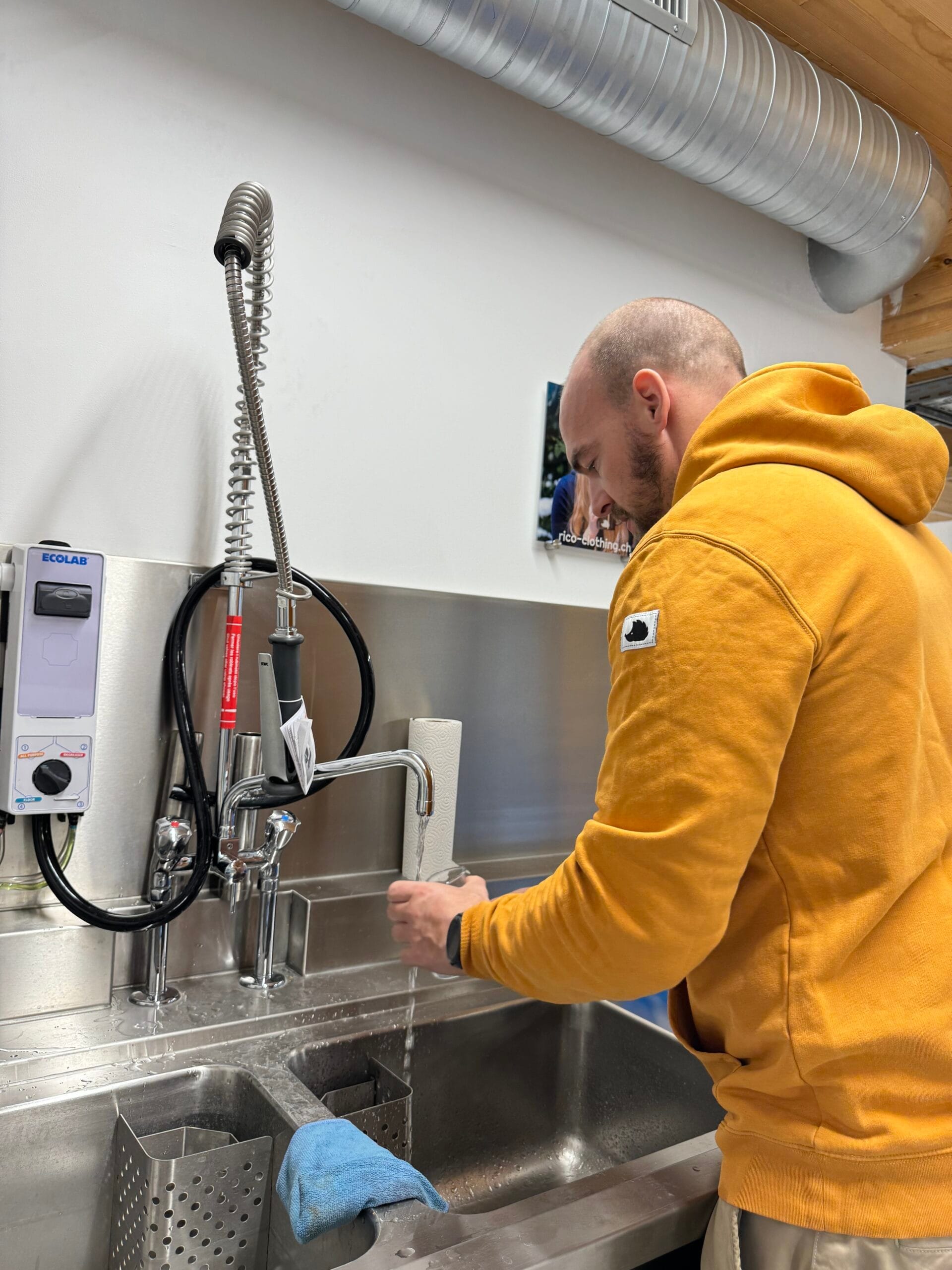 Ein Mann in einem orangefarbenen Hoodie spült an einem Doppelspülbecken aus Edelstahl in der Igel Station der Igel-Hilfe Schweiz.